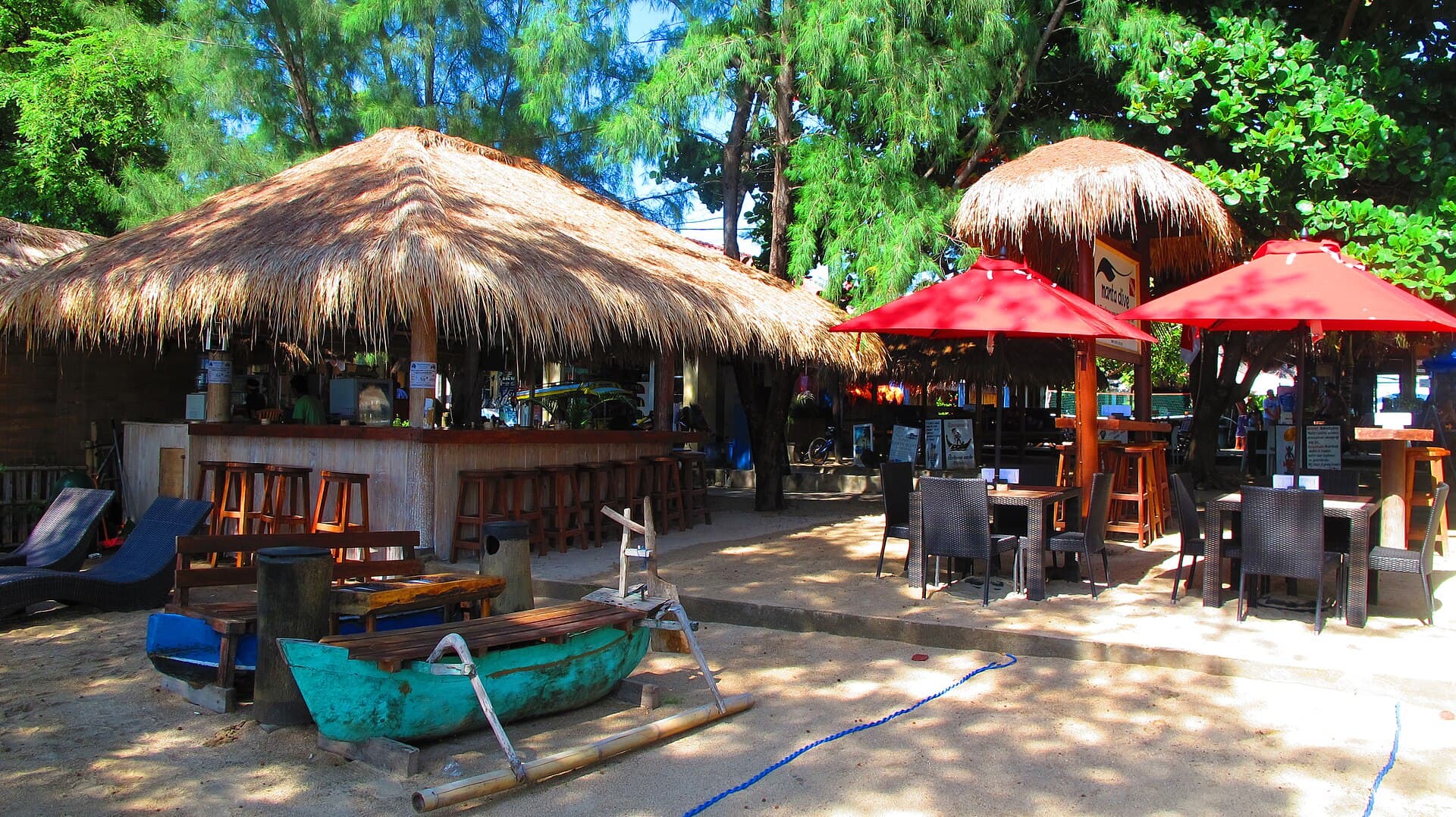 Beach bar by day with thatched umbrellas, red parasols and an outrigger boat on the sand