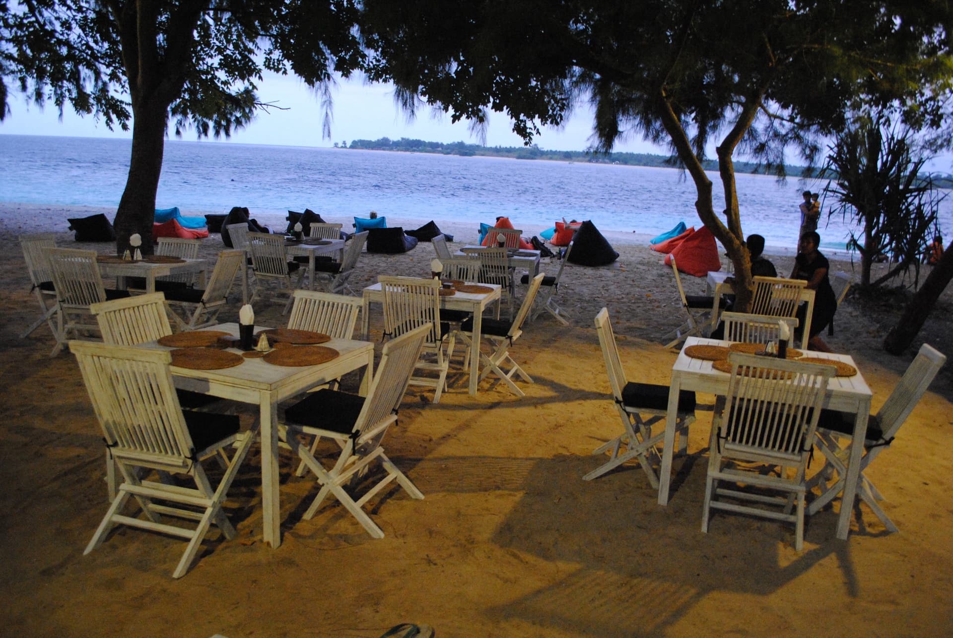 Beachfront café tables and chairs under trees at dusk