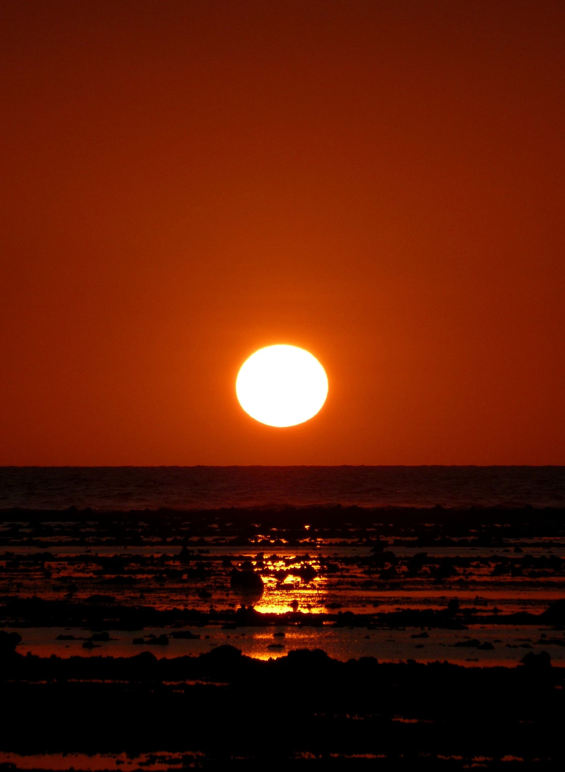 Orange sun setting low over the sea, with reef rocks in silhouette